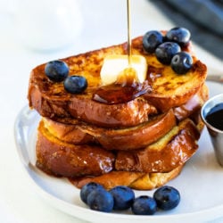 French Toast on a white plate with blueberries and syrup being poured on top.