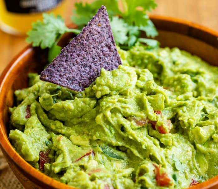 A tortilla chip dipping into a wooden bowl of guacamole with cilantro in the background.