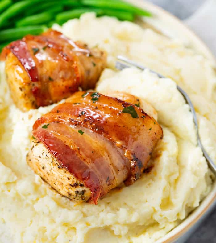 Bacon Wrapped Chicken on a pile of creamy mashed potatoes with green beans in the background.