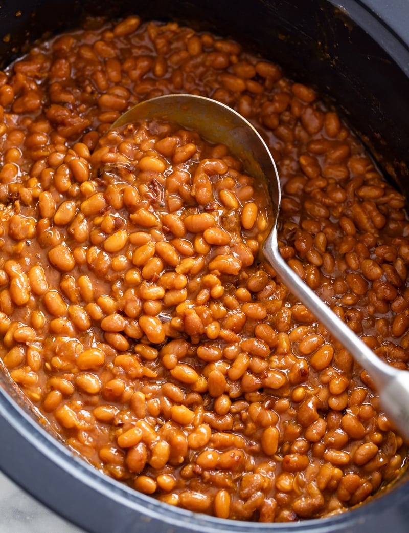 A crock pot filled with Baked Beans with a spoon in it.