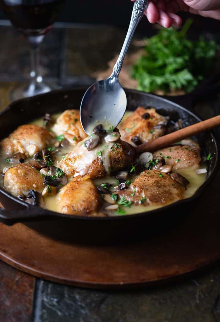 A cast iron skillet filled with golden chicken thighs with a spoon drizzling white wine sauce with mushrooms on top.