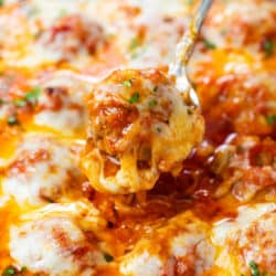 A spoon pulling up a cheesy baked meatball in marinara sauce from a casserole dish.