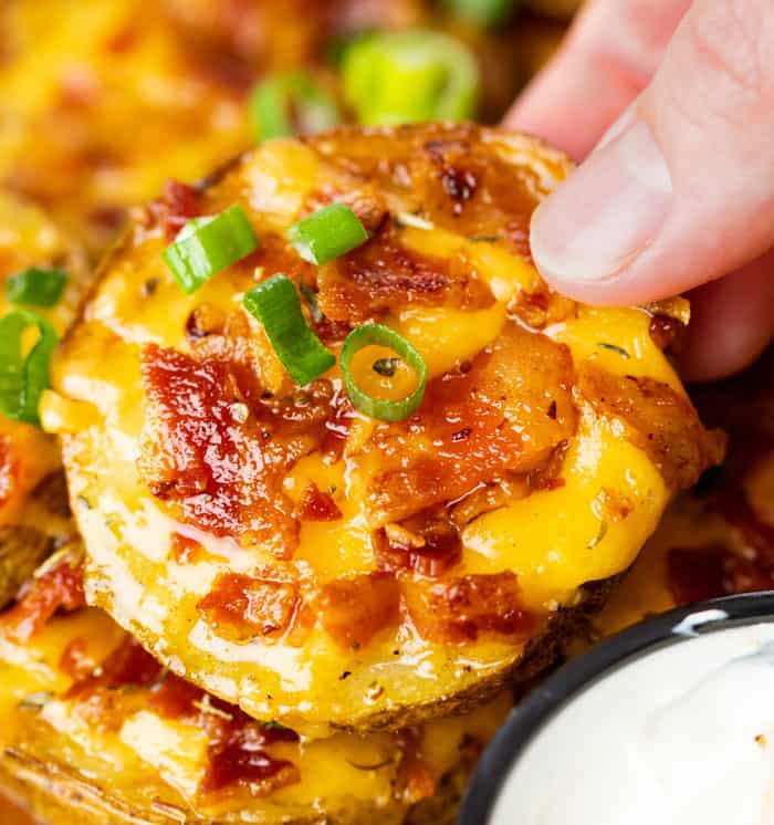 A hand holding a crispy slice of potato topped with melted cheese, bacon, and green onions.