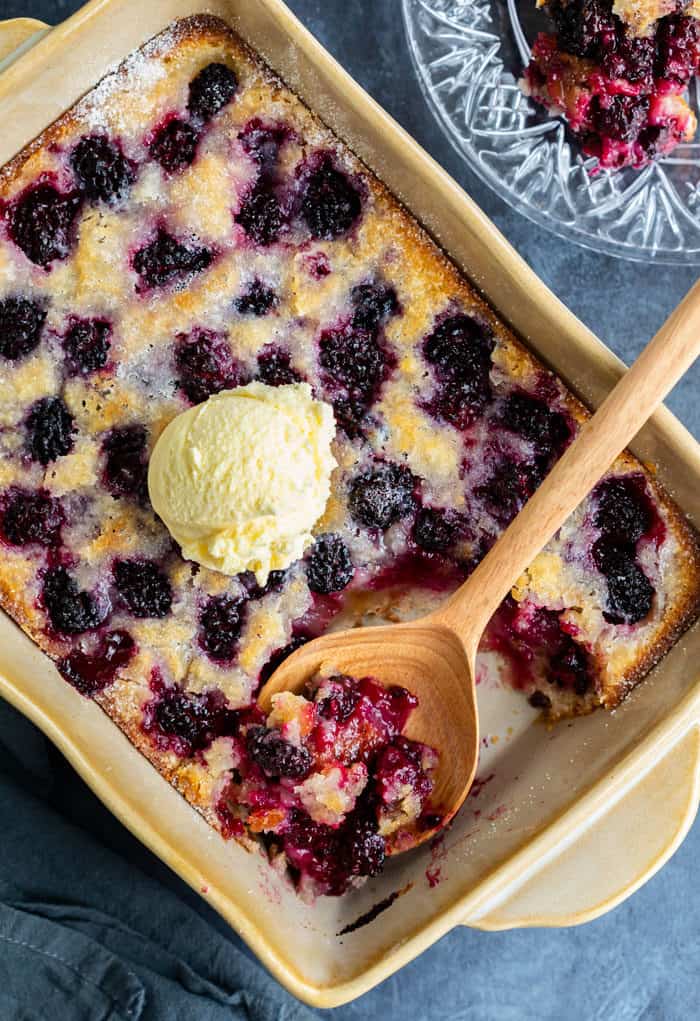 A casserole dish filled with Blackberry Cobbler with a wooden spoon and an ice cream scoop on top.