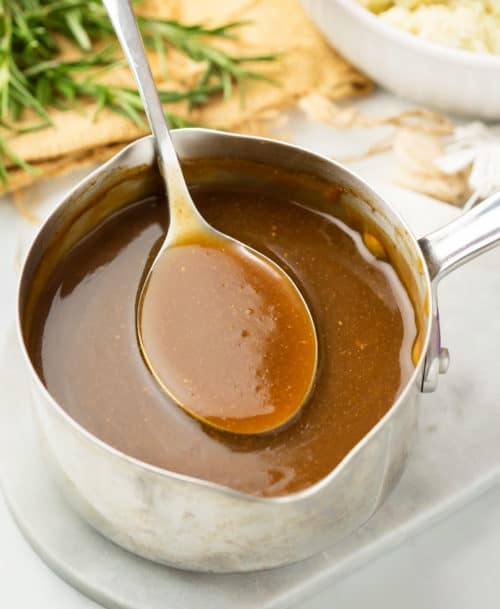 A pot of brown gravy with a spoon scooping it out with rosemary and mashed potatoes in the background.