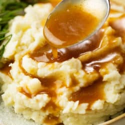 A spoon drizzling brown gravy over warm mashed potatoes.