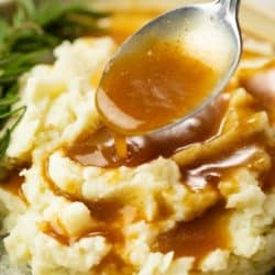 A pile of mashed potatoes on a plate with a spoon drizzling brown gravy on top.