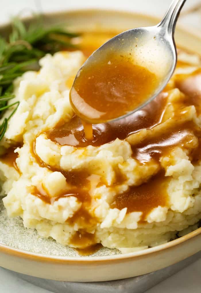 A spoon drizzling brown gravy over warm mashed potatoes.