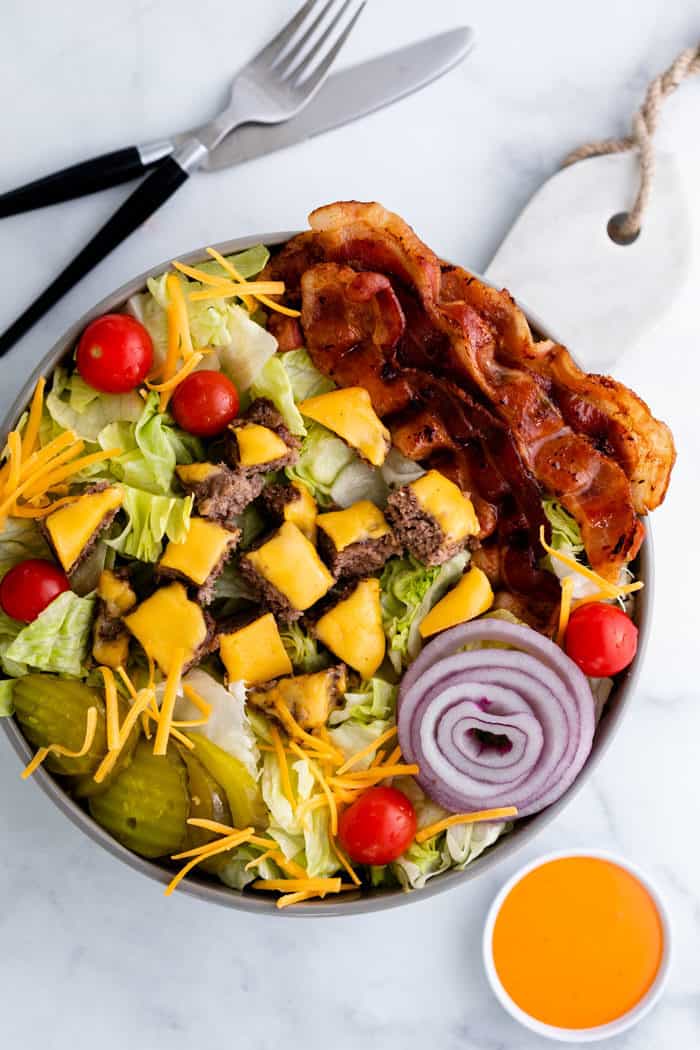 Overhead view of a big cheeseburger salad in a bowl on a white table with a fork, knife, and big mac sauce.