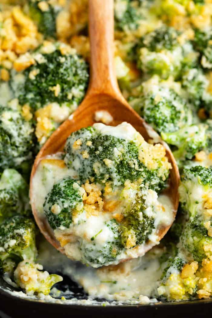 Close up image of a wooden spoon in a casserole dish filled with cheesy baked broccoli.