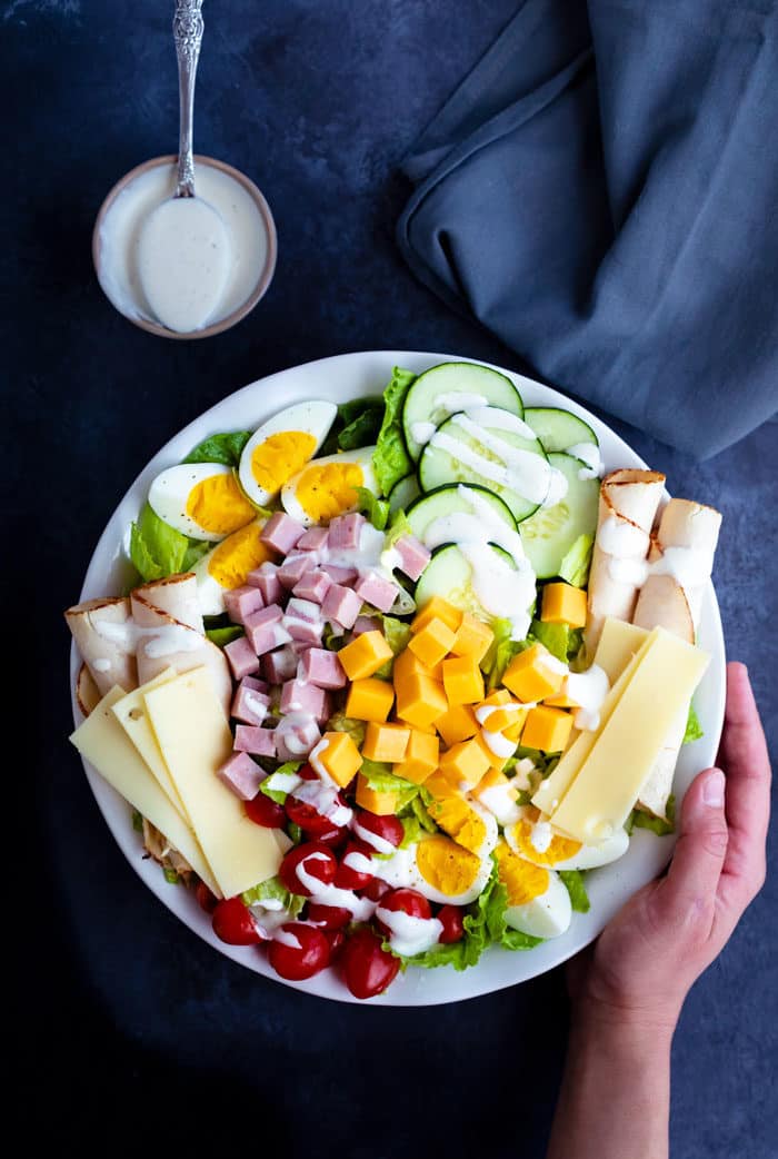 Overhead view of a white plate full of colorful chef's salad with a hand holding the side of the plate. The background is dark blue, there is a cup of ranch dressing to the side of the plate.
