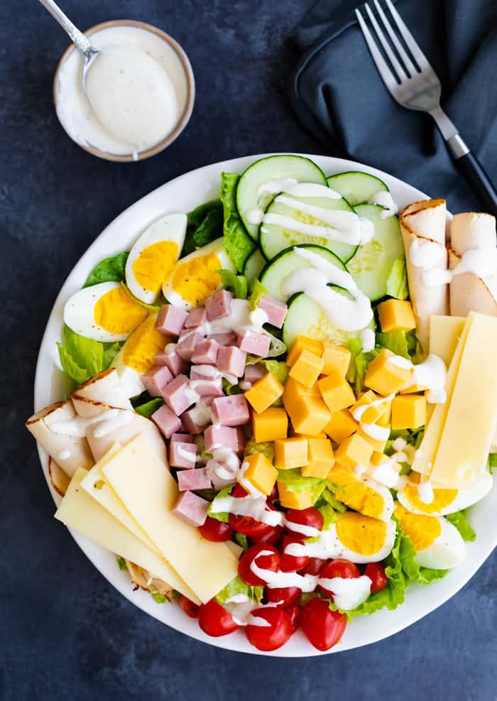 overhead view of a loaded, colorful chef salad on a white plate with a ramekin of ranch dressing and a fork next to it on a dark blue surface.