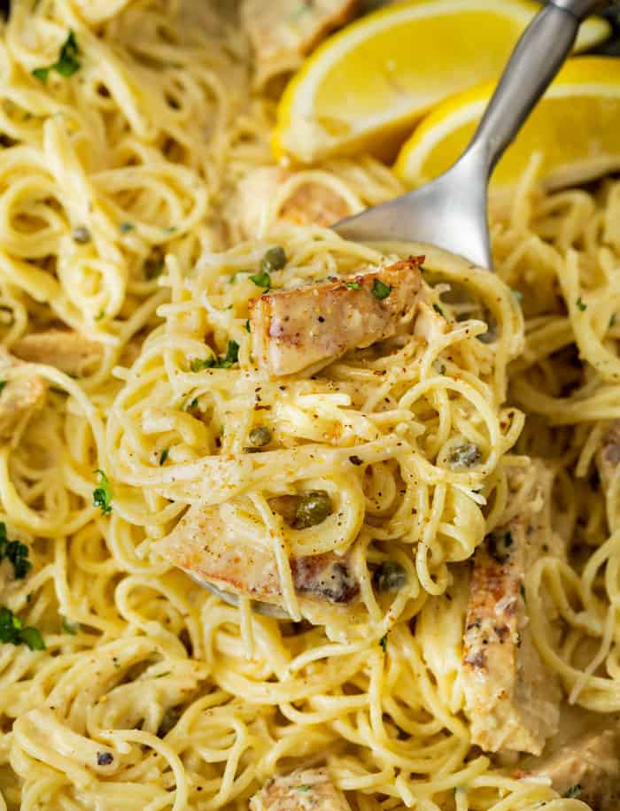 A spoon scooping up angel hair in a creamy Caesar sauce from a pot with capers and lemons.