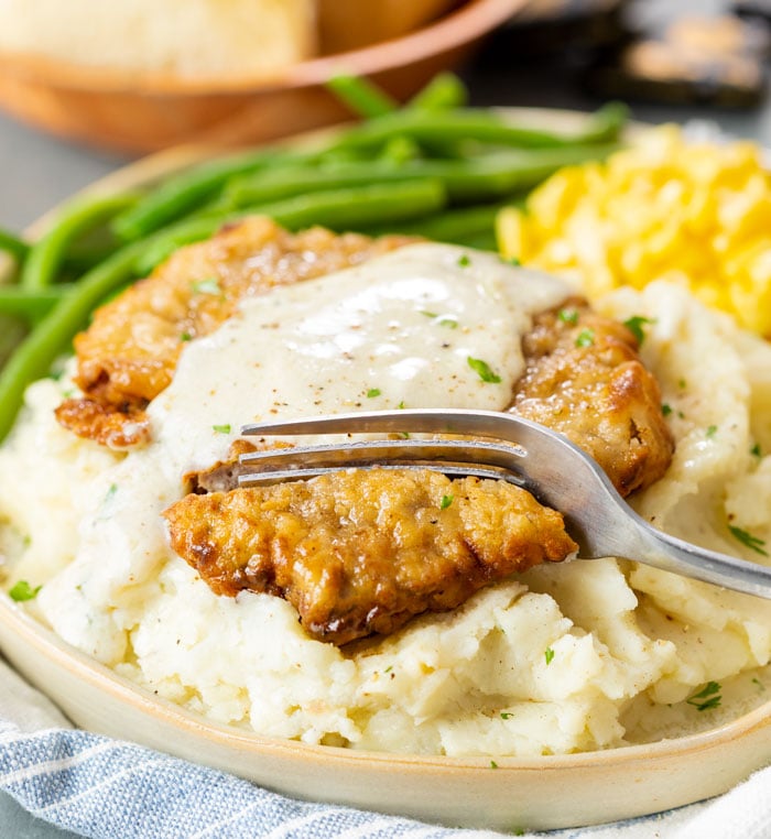 A fork cutting into a piece of chicken fried steak topped with white gravy over mashed potatoes.