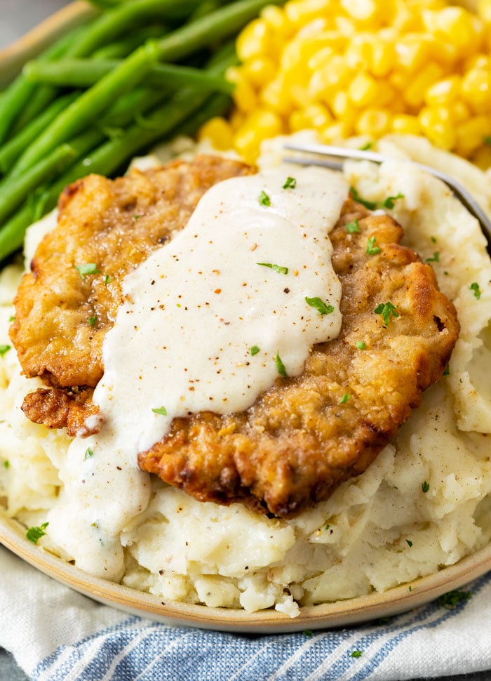A pile of mashed potatoes topped with chicken fried steak with white gravy on top and vegetables in the background.