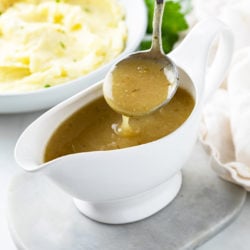 A ladle scooping up chicken gravy from a gravy boat with mashed potatoes in the background.
