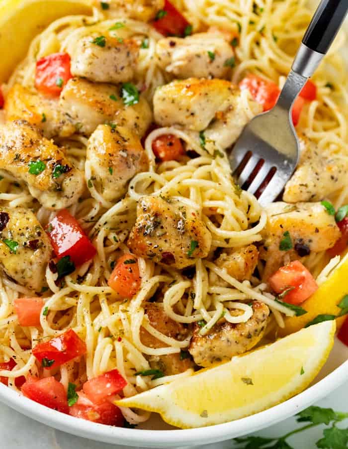 A fork in a bowl filled with chicken scampi with fresh tomatoes, parsley, and sliced lemon.