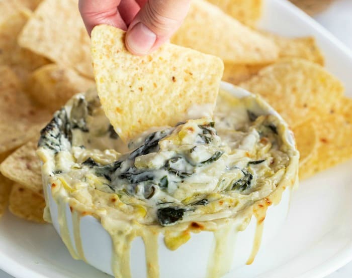 A hand dipping a tortilla dip into a white bowl filled with spinach and artichoke dip. Tortilla chips in the background.