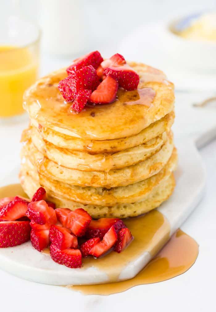 A tall stack of buttermilk pancakes topped with diced strawberries with a glass of orange juice in the background.