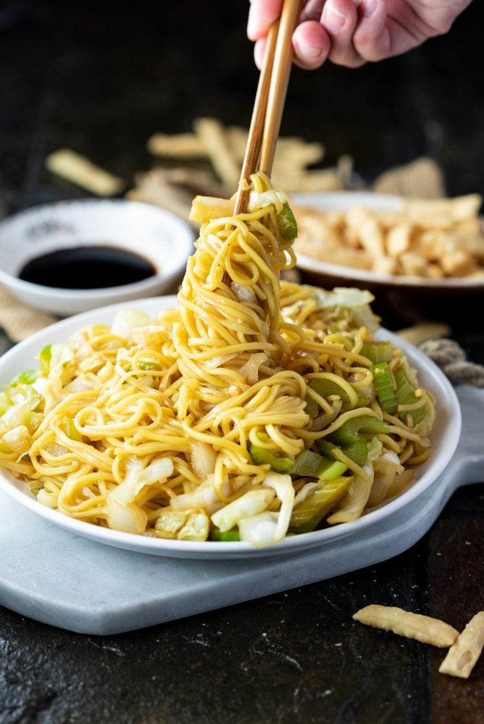 A white plate on a marble cutting board filled with chow mein and a hand holding chopsticks twisting up the noodles.