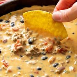 A hand dipping a tortilla chip into a skillet filled with Queso Dip with black beans, beef, and tomatoes.