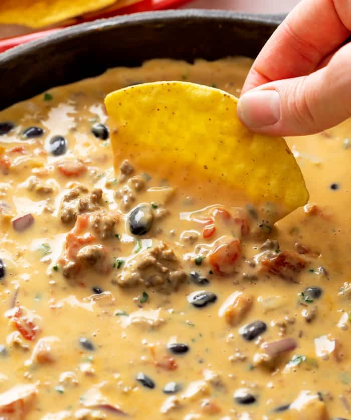 A hand dipping a tortilla chip into a skillet filled with Queso Dip with black beans, beef, and tomatoes.