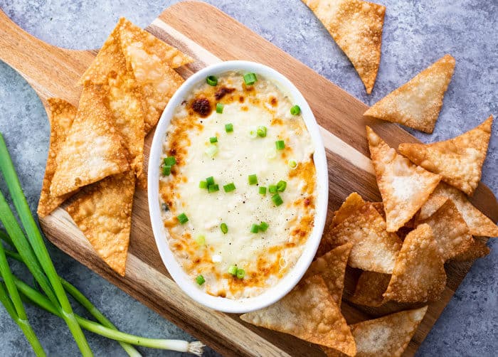 A white bowl of crab rangoon dip on a wooden cutting board with fried wontont chips scattered around and green onions.