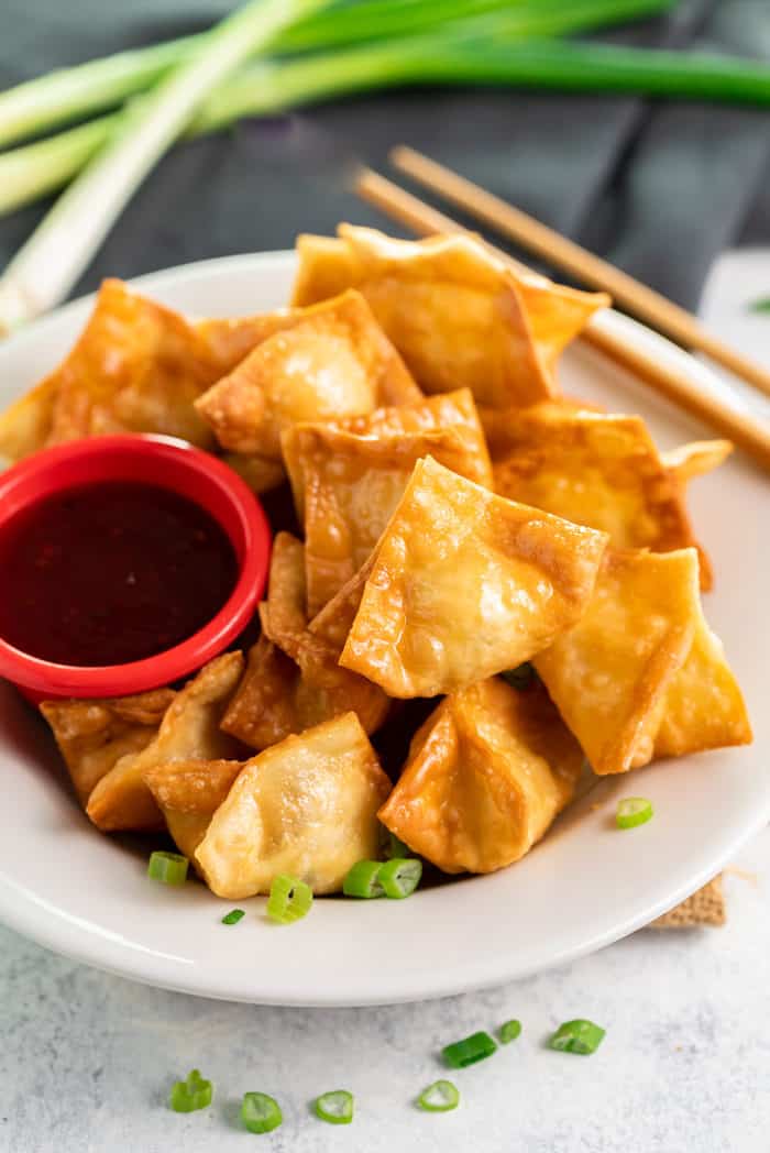 A white plate filled with fried crab rangoon and a red ramekin of sweet and sour sauce with chopsticks and green onions in background.