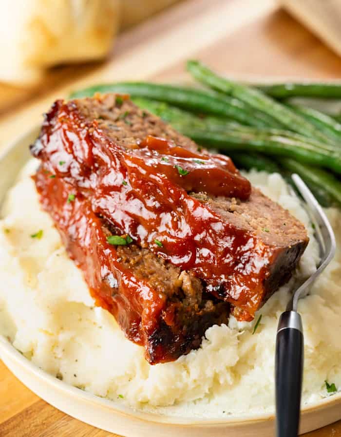 Cracker Barrel Meatloaf on a pile of mashed potatoes with green beans in the background.