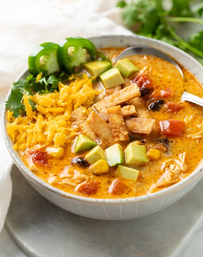 A bowl of Creamy Chicken Tortilla Soup topped with crispy tortillas, diced avocado, shredded cheese, jalapenos, and cilantro.
