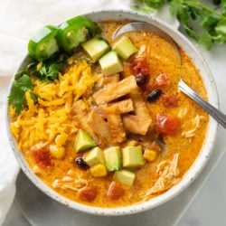 Overhead view of a white bowl with creamy chicken tortilla soup and garnishes like crispy tortillas, avocado, and shredded cheese.