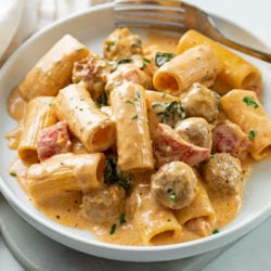 A white plate with creamy sausage pasta with roasted red peppers and rigatoni.