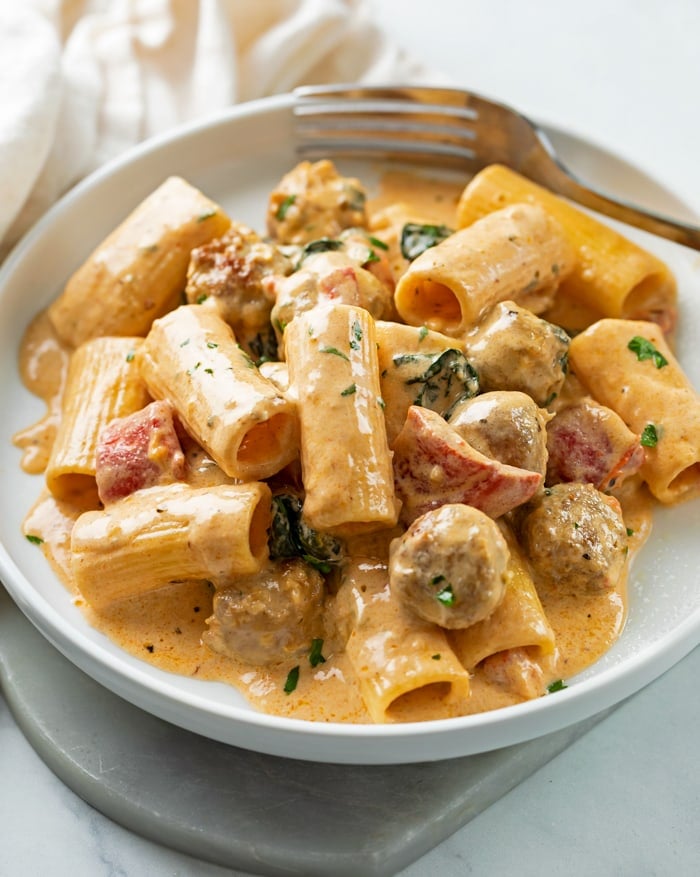 A white plate with creamy sausage pasta with roasted red peppers and rigatoni.