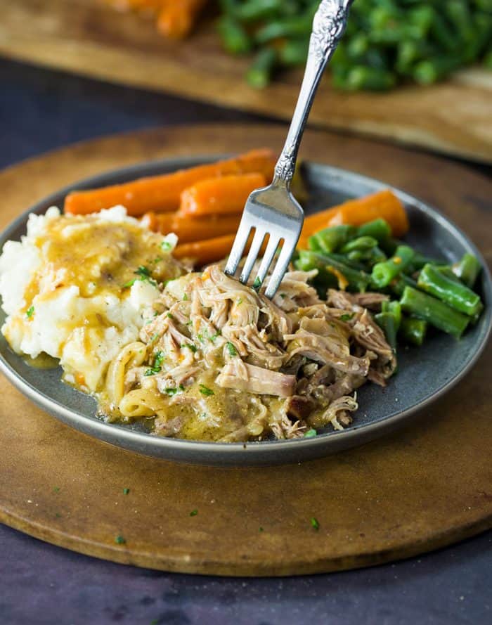 Shredded pork topped with parsley and gravy with a fork in it, with potatoes, carrots, and green beans in the background.