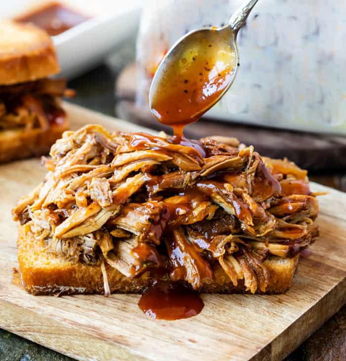 A spoon drizzling BBQ Sauce over pulled pork on a slice on bread.