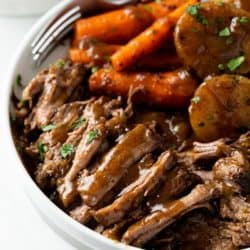A white plate with tender Crock Pot Roast with gravy and carrots and potatoes in the background.