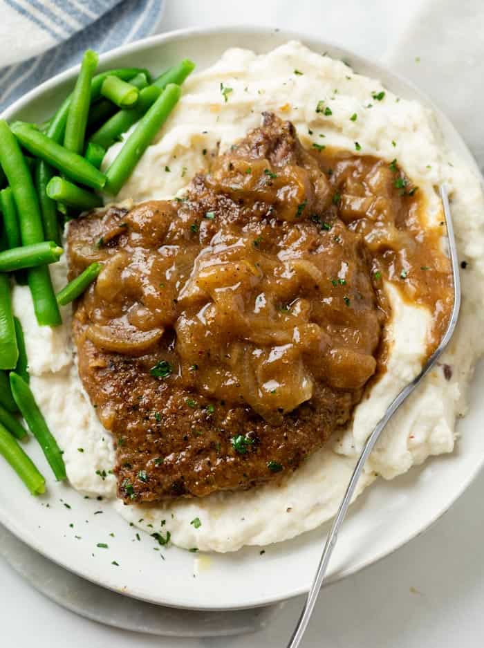 A pile of mashed potatoes topped with cubed steak with brown gravy and onions with green beans on the side.