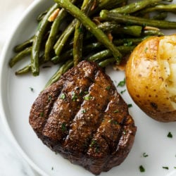 A filet mignon with steak rub on top next to roasted green beans and a baked potato.