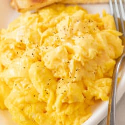 A white platter with fluffy scrambled eggs with a fork next to them and toast in the background.