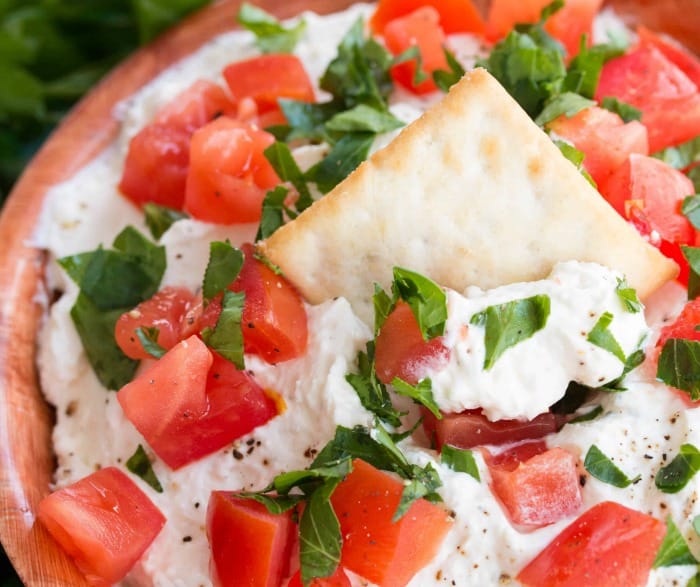 A close up view of a bowl of whipped garlic feta dip topped with diced tomatoes, parsley, and a chip in the middle.