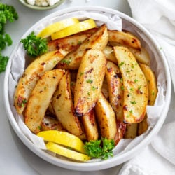 A white bowl of Greek Potatoes topped with parsley with lemon wedges on the side.