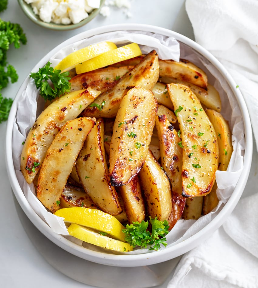 A white bowl of Greek Potatoes topped with parsley with lemon wedges on the side.