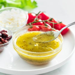 A spoon taking Greek Salad Dressing from a glass cup on a white plate.