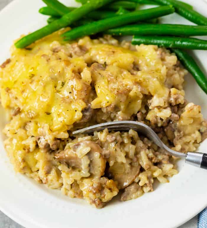 A white plate with cheesy ground beef and rice casserole with a fork. Green beans in the background.