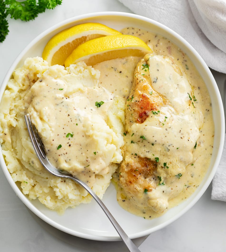 Creamy Herb Chicken on a white plate with mashed potatoes, cream sauce, and lemon wedges.