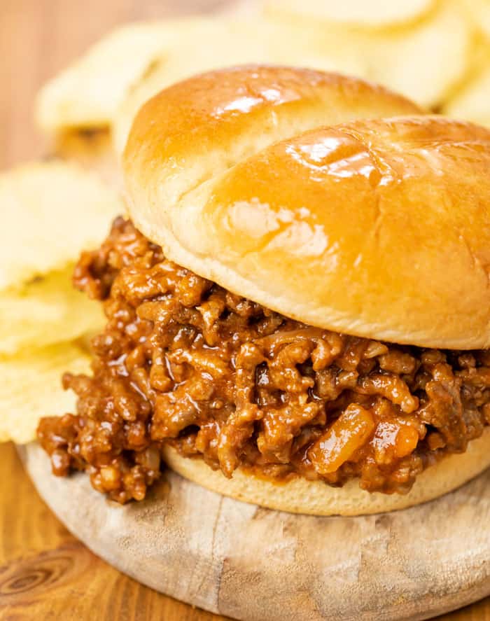 A hamburger bun filled with homemade sloppy joes on a wooden plate with chips in the background.