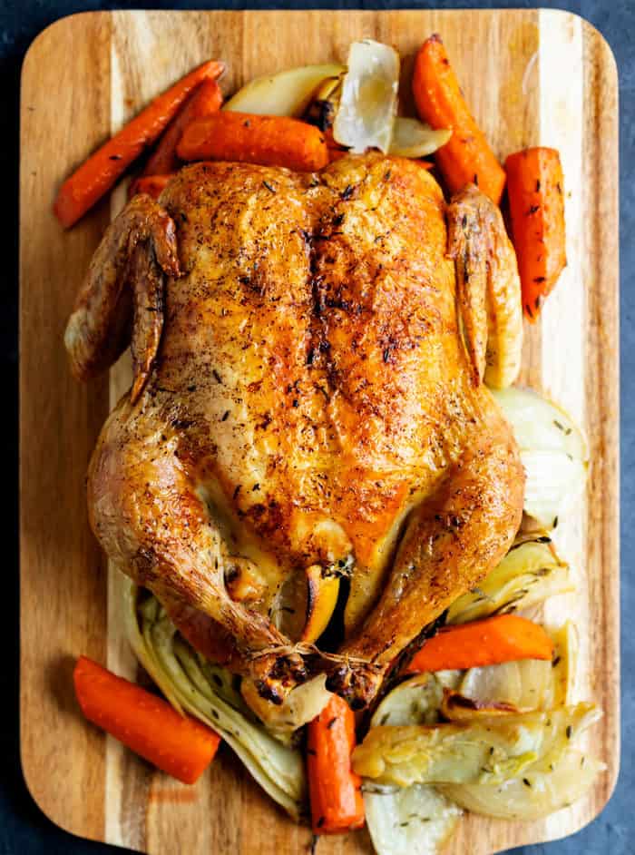 Overhead view of a cooked roasted chicken on a wooden cutting board surrounded by vegetables.