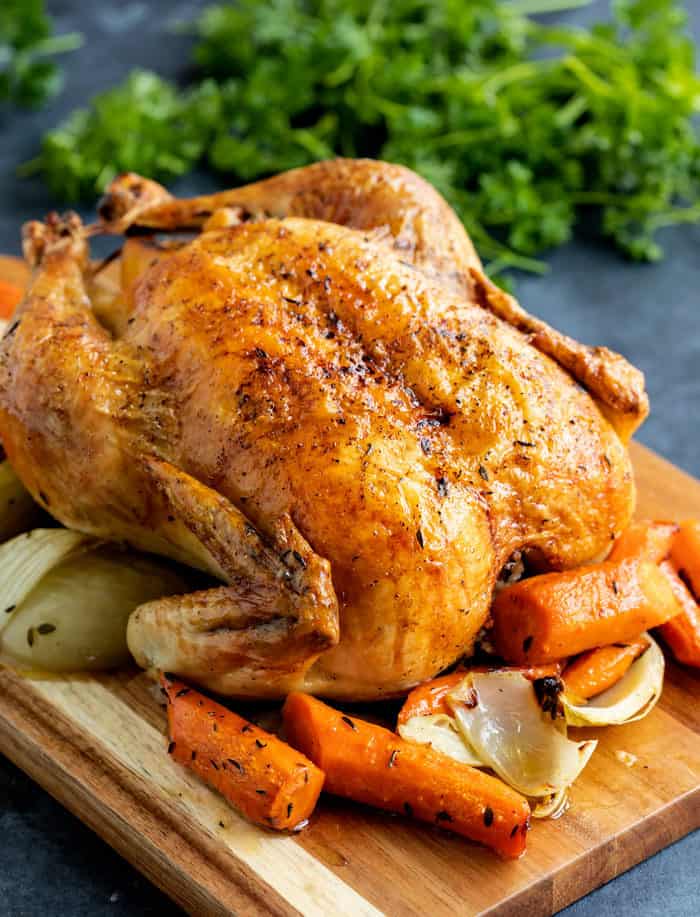 A brown roasted chicken on a wooden cutting board with carrots and onions, and parsley in the background.