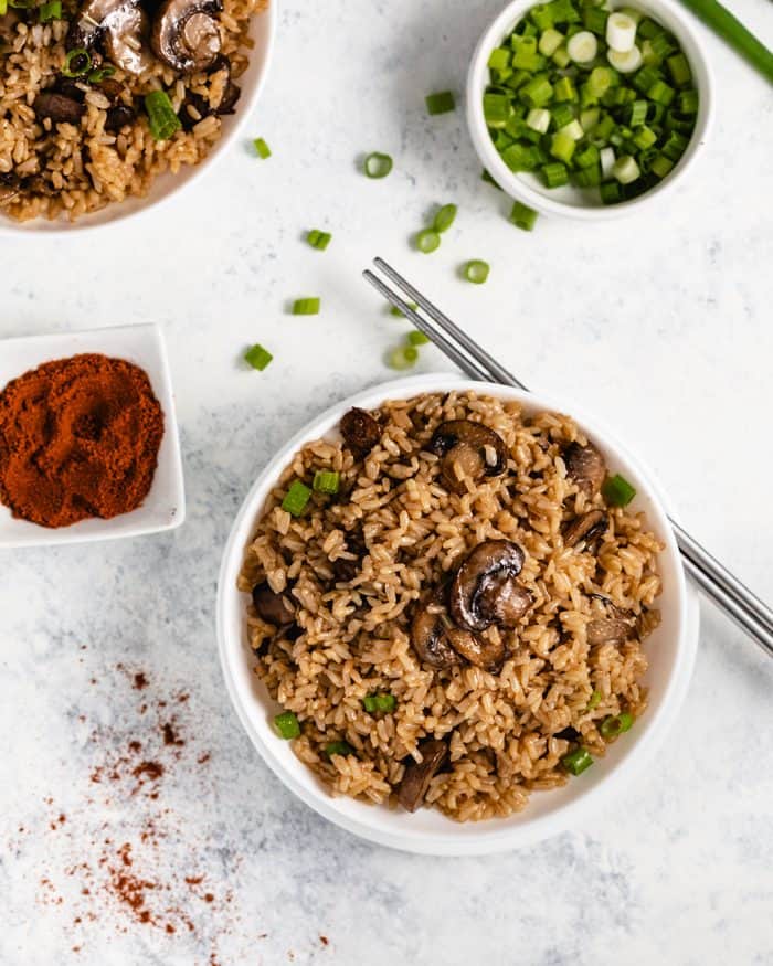 A white bowl with Brown Rice and mushrooms made in the Instant Pot next to green onions and paprika.