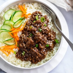 A white bowl of rice topped with Korean ground beef in sauce next to cucumbers and carrots.